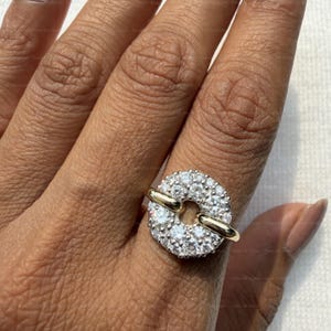 May include: A gold ring featuring a circular design encrusted with numerous small, sparkling white gemstones. The ring has a unique shape with a gold bar connecting the gem-studded circle.