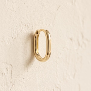 May include: A single polished gold-tone hoop earring with an elongated oval shape. The earring features a smooth, reflective metallic surface and a hinged closure mechanism at the top, showcasing a clean, continuous loop design with no additional embellishments.