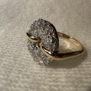 May include: A gold ring with a cluster of small, clear gemstones set in a circular design. The ring has a gold band and a gold accent that wraps around the gemstone cluster.