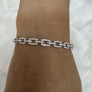 May include: A silver bracelet featuring a rectangular link design, adorned with small, sparkling gemstones. The bracelet is displayed on an arm, highlighting its delicate and elegant appearance. The gemstones reflect light, producing a dazzling effect.
