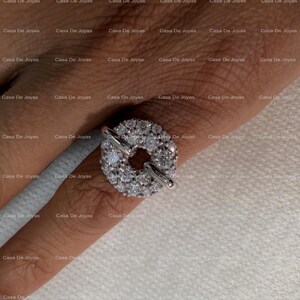May include: A silver ring with a circular, diamond-encrusted centerpiece and a clasp design. The ring is on a finger, showcasing its sparkling details. The ring is a piece of jewelry.