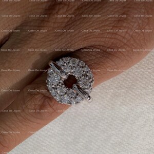 May include: A silver ring with a circular design encrusted with numerous small, clear gemstones. The ring features a bar closure. The background is a light skin tone.