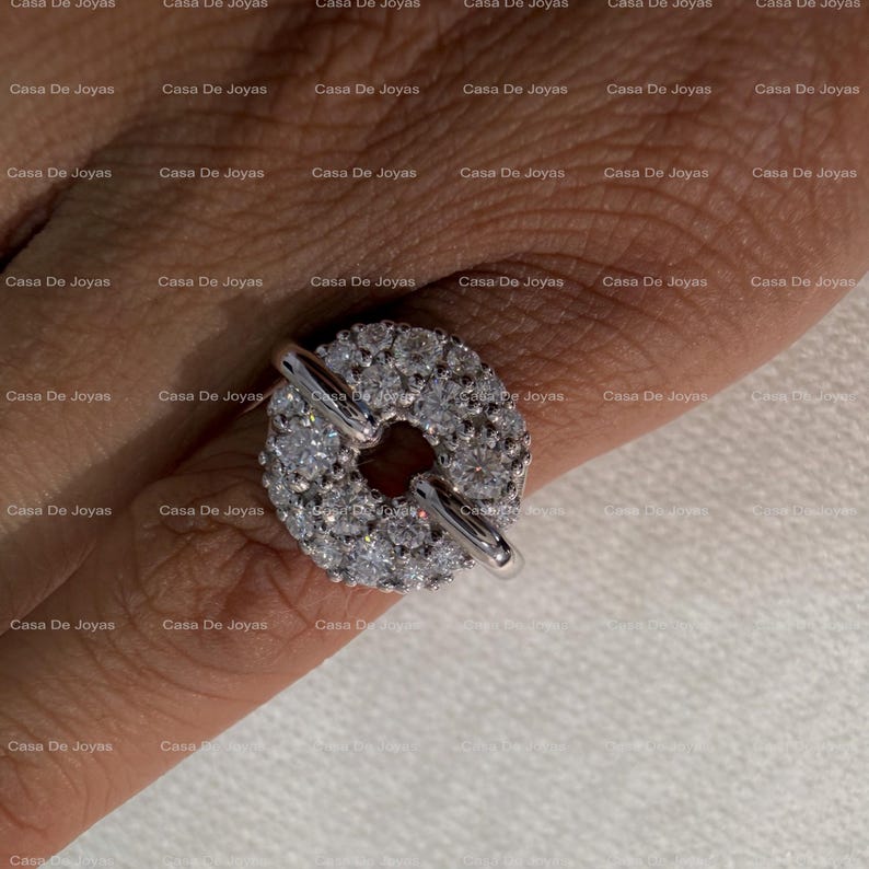 May include: A silver ring with a circular design encrusted with numerous small, clear gemstones. The ring features a bar closure. The background is a light skin tone.