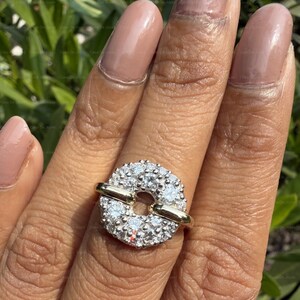May include: A ring featuring a circular design encrusted with numerous small, sparkling white stones. The ring has a gold band that passes through the center of the circle, creating a unique and eye-catching piece of jewelry.