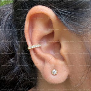 Könnte beinhalten: Ein goldener Ear Cuff mit einer Reihe kleiner, klarer, rund geschliffener Steine am äußeren Rand, platziert am oberen Knorpel, kombiniert mit einem einzelnen goldenen Ohrstecker mit einem klaren, rund geschliffenen Stein am Ohrläppchen.