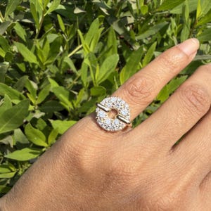 May include: A gold ring with a circular design, encrusted with small, clear gemstones. Two gold bars cross the center of the circle. The ring is worn on a finger, with a background of green foliage.