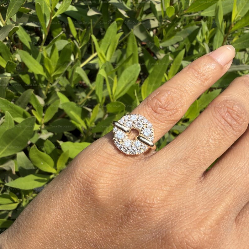 May include: A gold ring with a circular design, encrusted with small, clear gemstones. Two gold bars cross the center of the circle. The ring is worn on a finger, with a background of green foliage.