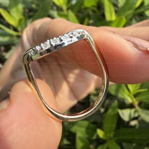May include: A gold-toned ring with a curved design, featuring a row of small, clear gemstones. The ring is held against a backdrop of green foliage, highlighting its polished surface and intricate details. The gemstones sparkle.