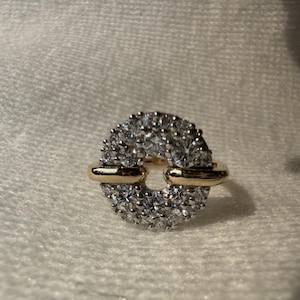 May include: A gold and silver ring with a circular design. The ring features a band of small, sparkling stones set in a silver metal, with gold accents on the sides. The ring is set against a white background.
