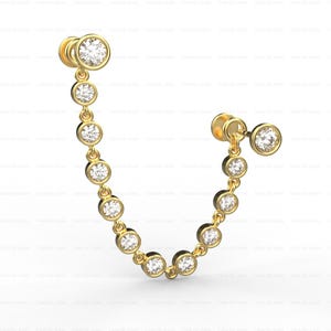 May include: A pair of gold-coloured double-piercing earrings. Each earring features a large, round, clear gemstone at the top, with a chain of smaller, round gemstones suspended below. The gemstones are set in a bezel setting.