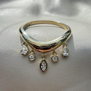 Pode incluir: Um anel de ouro com uma banda curva e cinco pingentes pendurados. Os dois pingentes externos têm formato de gota, os dois internos são redondos e o pingente central tem formato de marquise. Cada pingente possui uma pedra transparente facetada.