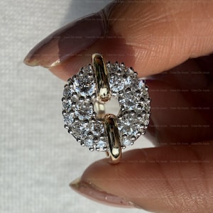 May include: A gold-toned ring with a circular design, encrusted with numerous small, clear gemstones. The ring features a central opening and two gold bars connecting the circular elements. The ring is held between fingers.