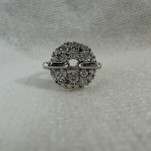 May include: A silver ring with a circular design, encrusted with numerous small, clear gemstones. The ring features two horizontal silver bars that connect to the circular gemstone cluster, creating a unique and elegant look. The ring is set against a white background.