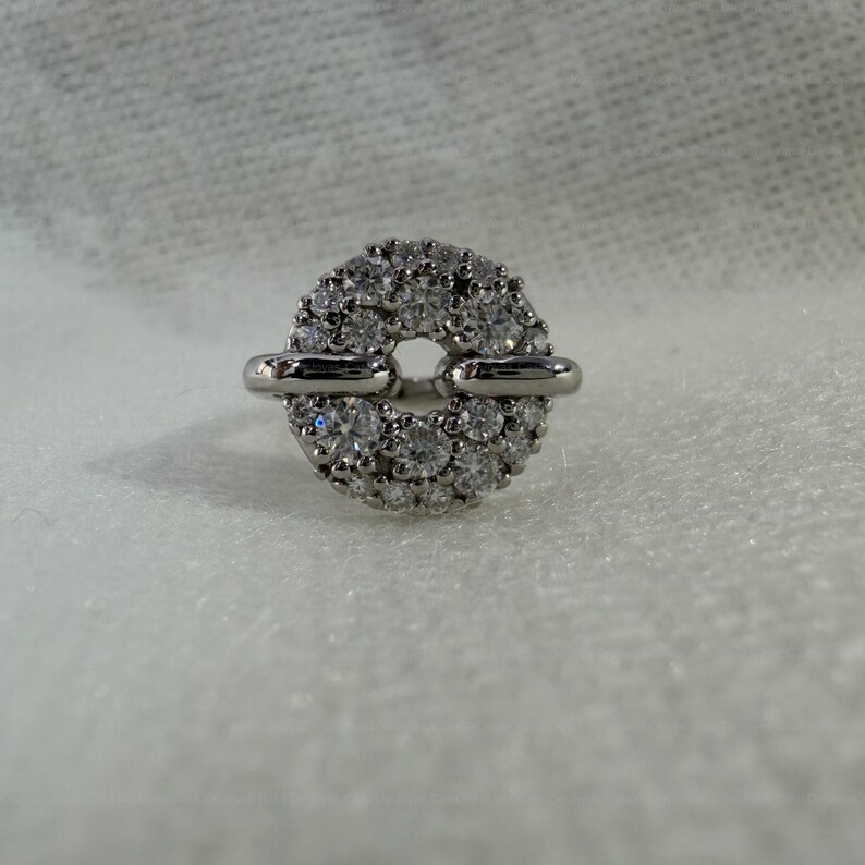 May include: A silver ring with a circular design, encrusted with numerous small, clear gemstones. The ring features two horizontal silver bars that connect to the circular gemstone cluster, creating a unique and elegant look. The ring is set against a white background.