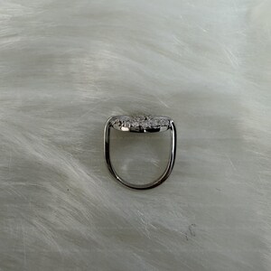 May include: A silver ring with a heart-shaped design, adorned with small, sparkling stones. The ring's band is a simple, polished silver, and the heart is set with multiple clear gemstones. The ring is displayed on a white, textured surface.
