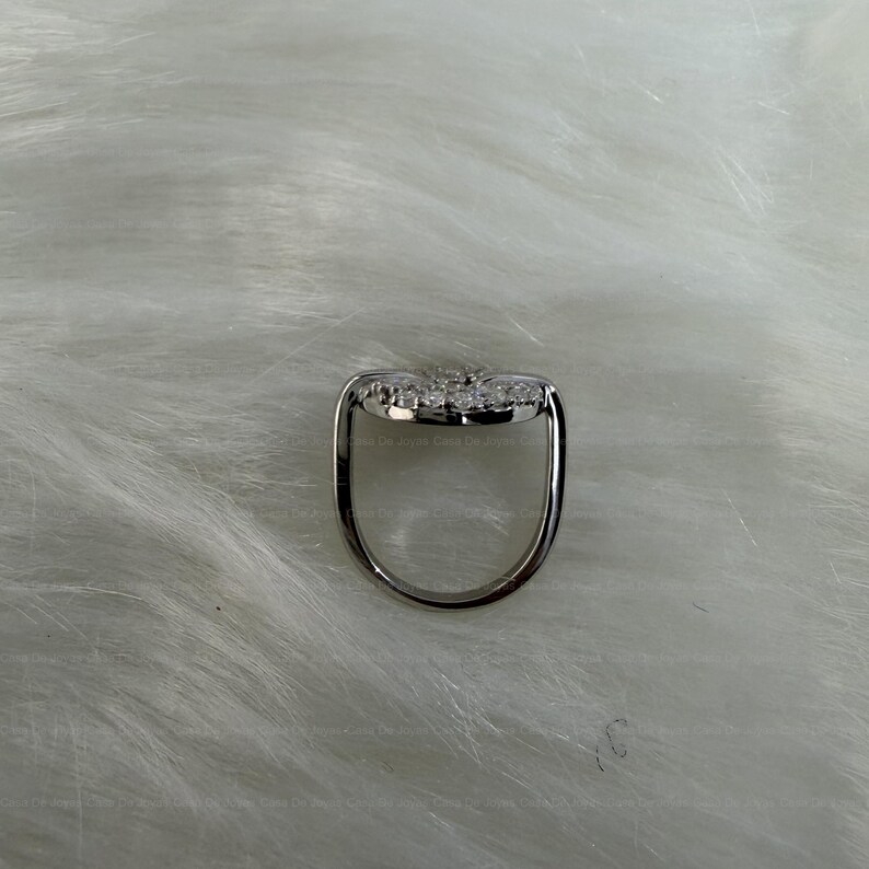 May include: A silver ring with a heart-shaped design, adorned with small, sparkling stones. The ring's band is a simple, polished silver, and the heart is set with multiple clear gemstones. The ring is displayed on a white, textured surface.
