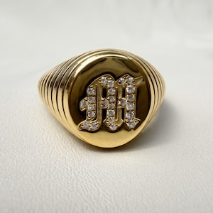 May include: A gold signet ring featuring a prominent, raised letter M in an old English font. The letter is encrusted with small, clear, sparkling stones. The ring band has a distinct ribbed or grooved texture that wraps around the sides.
