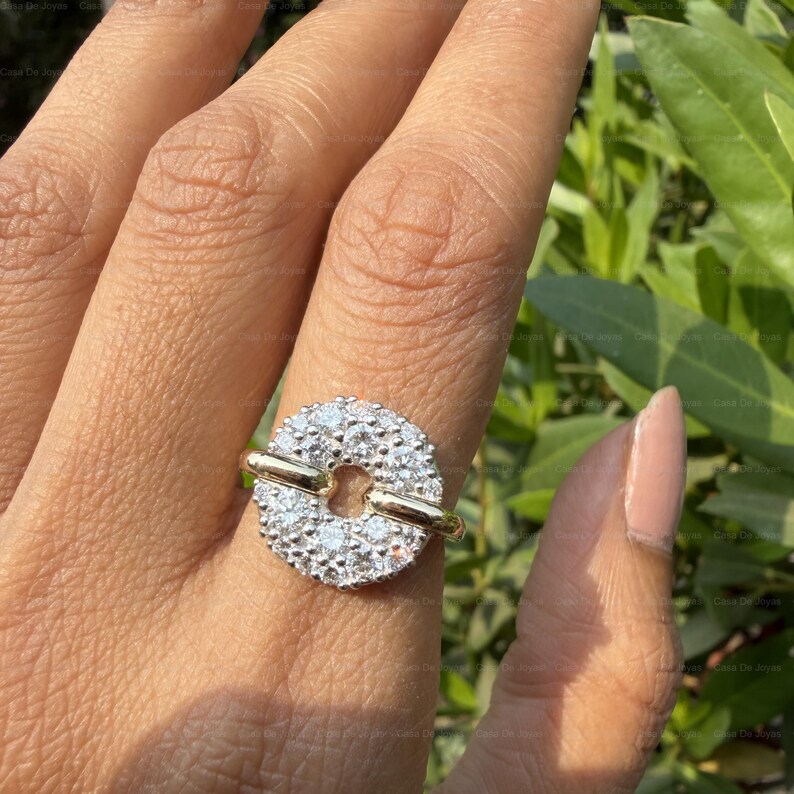May include: A gold ring with a circular, diamond-studded centerpiece. The ring features a unique design with a horizontal gold bar across the center of the circle. The ring is shown on a person's finger.