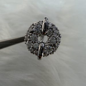 May include: A close-up of a silver-toned ring with a circular design. The ring is encrusted with numerous small, clear gemstones, creating a sparkling effect. Two vertical silver bars are attached to the ring. The ring is held by a silver tool.
