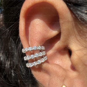 May include: Three silver ear cuffs, each adorned with a row of sparkling, clear gemstones. The ear cuffs are worn on the ear, creating a layered effect. The jewelry is set against the backdrop of the ear and skin.