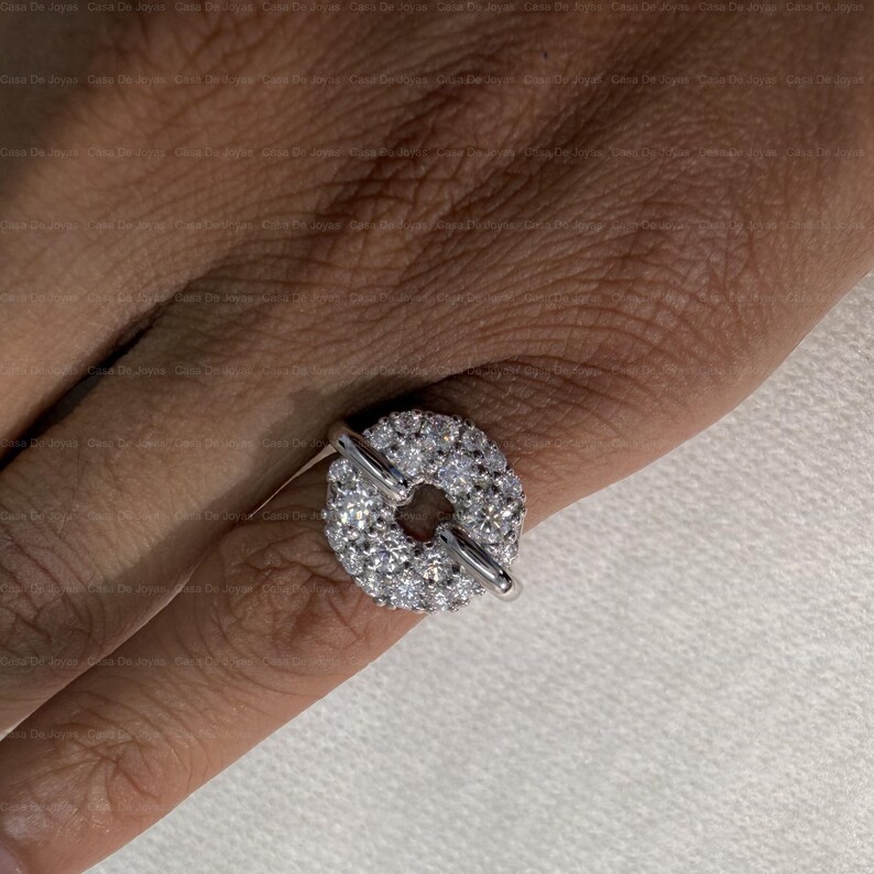 May include: A silver ring featuring a circular design encrusted with numerous small, clear gemstones. The ring has a unique toggle closure, with a bar that slides through a loop. The ring is shown on a finger.