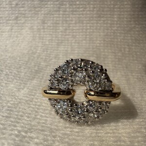 May include: A diamond ring with a circular design. The ring features a gold band with a cluster of small diamonds around the outer edge. The center of the ring is open, creating a unique and elegant look.