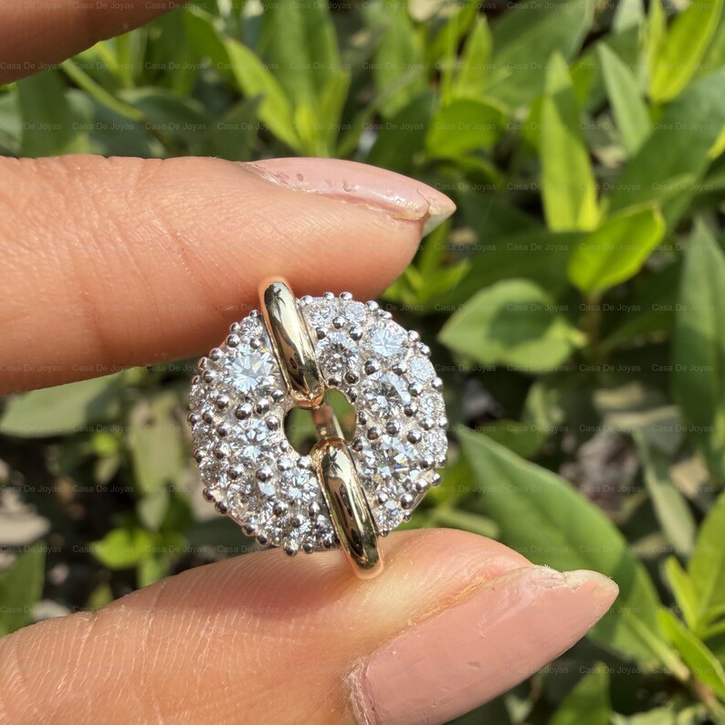 May include: A round, gold-toned earring with a cluster of small, clear gemstones. The earring has a central opening and a gold bar that connects the two halves. The background is blurred green foliage.