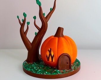 May include: A 3D-printed Halloween decoration featuring an orange pumpkin house with a brown door and window. A brown tree with green leaves stands beside the pumpkin on a base with green grass. The decoration is designed for Halloween.