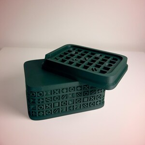 May include: A dark green box and lid. The lid has a grid pattern, and the box's sides feature a repeating geometric design. The box and lid are made of a solid material. The box is rectangular.