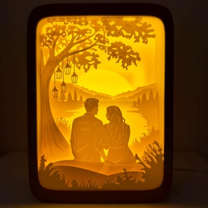 May include: A warm, illuminated paper cut light box featuring a couple seated under a tree, overlooking a lake and mountains at sunset. The scene is lit with a soft, yellow glow, creating a romantic ambiance. The frame is a dark brown color.