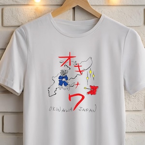 Può includere: T-shirt bianca con un design grafico colorato raffigurante una mappa di Okinawa, in Giappone, con caratteri giapponesi e il numero 7. Il design include un personaggio dei cartoni animati e il testo "OKINAWA JAPAN".