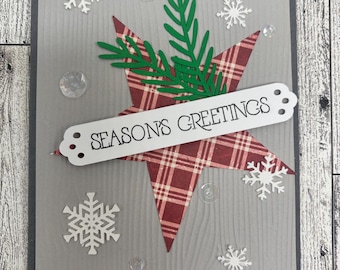 May include: A handmade greeting card with a gray background and a dark gray border. The card features a red plaid star, green sprigs, white snowflakes, and the text "SEASON'S GREETINGS". Clear, iridescent sequins are scattered around the design.