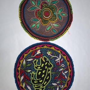 Könnte beinhalten: Zwei runde, gestickte Aufnäher. Der obere Aufnäher hat einen braunen Hintergrund mit einem floralen Design in Grün, Gelb und Rot. Der untere Aufnäher hat ein schwarzes Froschdesign auf blauem Hintergrund, umgeben von farbenfrohen Blumenstickereien.