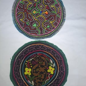 Könnte beinhalten: Zwei runde, bestickte Textilstücke. Das obere Stück hat ein komplexes geometrisches Design in Rot, Gelb, Grün und Blau auf schwarzem Hintergrund. Das untere Stück zeigt eine braune Schlange mit gelben Blumen und grünen Blättern auf schwarzem Hintergrund.