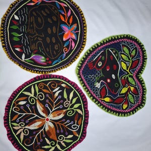 Könnte beinhalten: Drei bestickte schwarze Stoffstücke mit farbenfrohen Blumen- und Tiermotiven. Eines ist rund mit einer jaguarartigen Figur, ein anderes herzförmig mit einem Frosch, und das dritte rund mit einem Blumenmuster. Jedes hat einen dekorativen Rand.