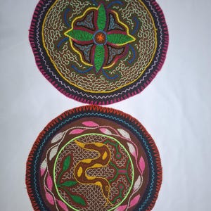Könnte beinhalten: Zwei runde, bestickte Textilstücke. Eines zeigt ein zentrales grünes Blumendesign mit orangefarbenen Akzenten, umgeben von braunen und weißen Nähten. Das andere hat ein gelbes Schlangendesign mit grünen Blättern und rosa Akzenten. Beide haben dunkelbraune und rosa Ränder.