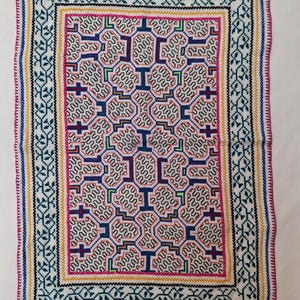 Op de afbeelding: Rechthoekig textiel met ingewikkelde kruissteekborduurwerk. Het ontwerp heeft een centraal patroon van onderling verbonden vormen in wit, blauw en rood, omlijst door randen van groen, geel en blauw. De randen zijn afgewerkt met een roze gestikte rand.