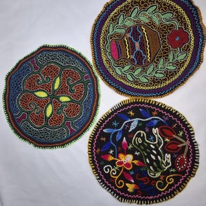 Könnte beinhalten: Drei runde, bestickte Textilstücke mit aufwendigen Designs. Eines zeigt ein zentrales Blumenmuster, ein anderes eine Vase mit Blättern und das dritte einen Frosch mit Blumen. Jedes hat einen farbenfrohen Rand und detaillierte Stickereien.