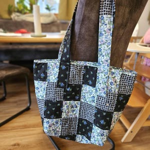 Könnte beinhalten: Eine Patchwork-Tragetasche mit Blumen- und Tupfenmuster. Die Tasche hat eine Kombination aus schwarzen, weißen und blauen Stoffen mit einem gesteppten Design. Die Griffe sind aus dem gleichen Blumenstoff gefertigt.