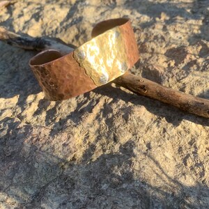 Peut inclure: Un bracelet manchette en cuivre martelé avec une section centrale dorée et des rivets argentés. Le bracelet repose sur une surface rocheuse texturée, avec un bâton en bois à proximité. Le bracelet est de conception large et ouverte.