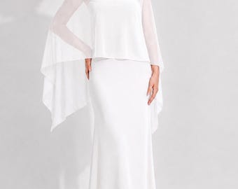 May include: A floor-length white gown featuring a strapless bodice and a sheer, lightweight overlay with long, flowing cape-style sleeves that extend from the shoulders. The dress has a clean, solid white finish and a straight, column-like silhouette.