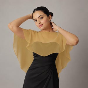 May include: A sheer gold capelet featuring a wide, flowing silhouette with ruffled edges, worn over a sleeveless black dress. The capelet is made of a lightweight, translucent fabric that drapes over the shoulders and upper torso.