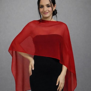 Puede incluir: Un chal rojo vibrante y transparente drapeado sobre los hombros, confeccionado con una tela ligera y translúcida que cae en pliegues suaves. El chal se lleva sobre una prenda negra lisa, creando un contraste marcado con su tono rojo.