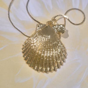 May include: A gold-toned metal pendant shaped like a scallop shell, featuring a textured, ribbed surface with a metallic finish. The pendant is attached to a thin, matching gold-toned chain that loops behind the shell, creating a cohesive piece.