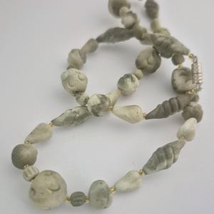 May include: A necklace featuring a series of irregularly shaped, matte-finish beads in shades of grey, white, and muted green. The beads vary in form, including rounded, teardrop, and ribbed shapes, separated by small, clear, translucent spacers.