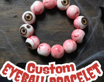 Puede incluir: Un brazalete de ojo personalizado con cuentas rosas y blancas con venas rojas e iris marrones. El brazalete se muestra sobre una superficie de madera con decoraciones de Halloween. El texto "Custom Eyeball Bracelet" está en la imagen.