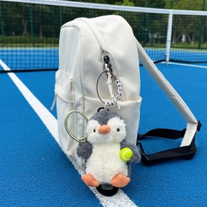 Puede incluir: Mochila color crema con un bolsillo frontal y una correa negra. Un llavero de pingüino de peluche con una raqueta y una pelota de tenis cuelga de la cremallera. El fondo es una cancha de tenis azul.