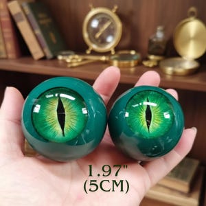 Könnte beinhalten: Zwei grüne Glaskugeln, jede mit einem detaillierten Reptilienaugen-Design, schwarzer Pupille und grüner Iris. Die Kugeln haben einen Durchmesser von etwa 5 cm. Der Hintergrund umfasst antike Gegenstände.