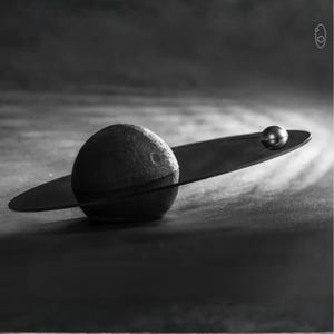 Può includere: Scultura decorativa in bianco e nero raffigurante un pianeta con una sfera strutturata, un anello circostante e una piccola sfera metallica. La scultura è ambientata su uno sfondo scuro e strutturato, creando un contrasto drammatico.