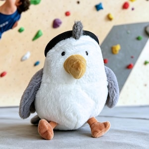 以下が含まれることがあります： 白い体、灰色の翼、黒い帽子をした、ぬいぐるみのアオサギのおもちゃ。アオサギは黄色のくちばしとオレンジ色の足を持っています。おもちゃは灰色の表面に座っており、背景にはクライミングウォールがあります。